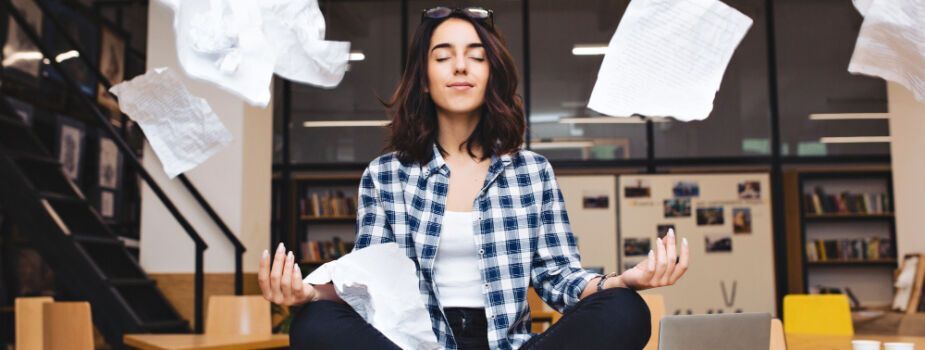 Jeune femme assise en tailleur sur son bureau avec ses dossiers qui volent pour symboliser la réduction de sa charge mentale.